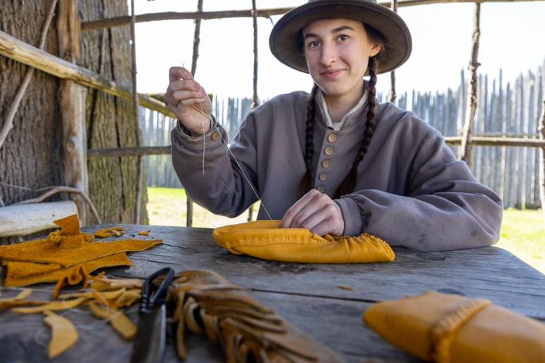 A student interpreter in costume making a moccasin on the historic site